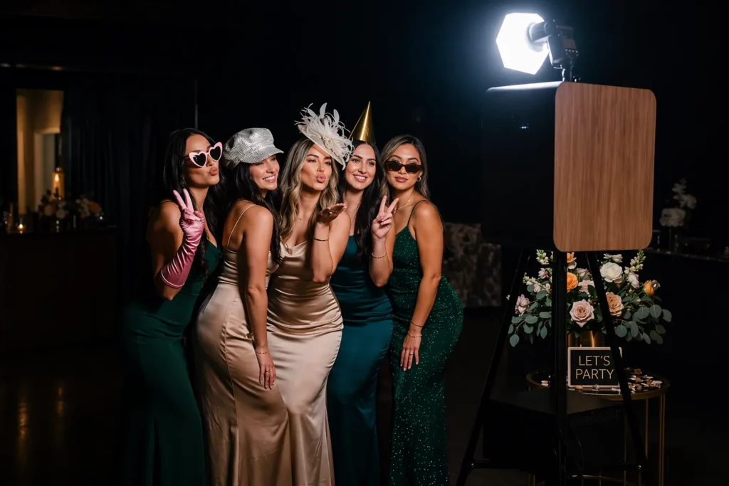 Un groupe de 5 filles posant devant un photobooth avec des chapeaux et lunettes fun